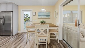 A dining area with a wooden table, white chairs, a hanging light fixture, a refrigerator, and a sliding glass door leading to a balcony with a view of buildings.