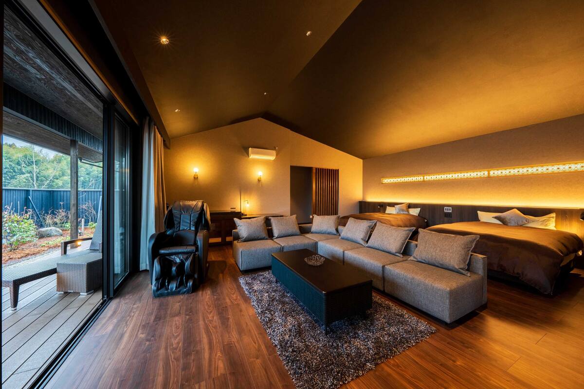 A modern living room with a sectional sofa, a black leather chair, a coffee table, and a large window offering a view of the outdoors.