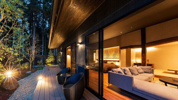 Eine Terrasse im Freien mit einer Couch und Stühlen, Holzboden und einer Glaswand, die in einen Raum mit einem Bett und einer Lampe führt.