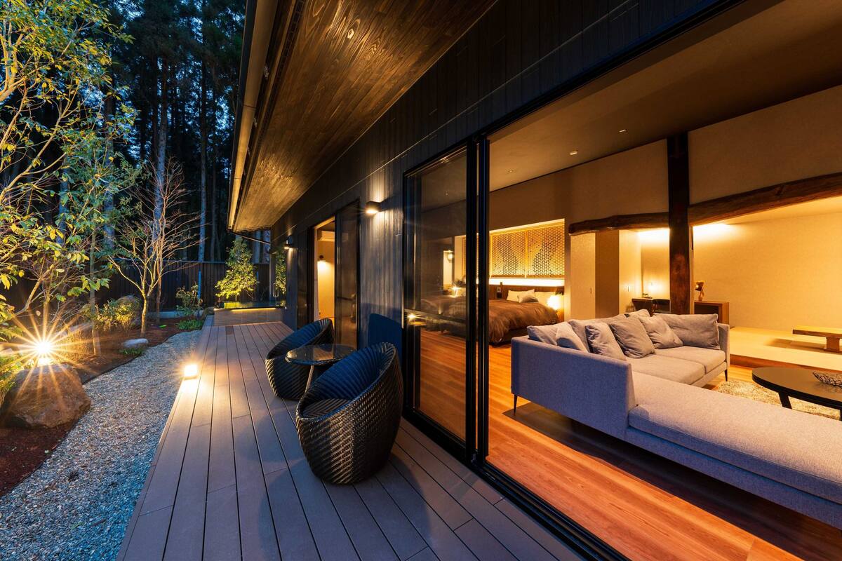 An outdoor deck with a sofa and chairs, a wooden floor, and a glass wall leading to a room with a bed and a lamp.