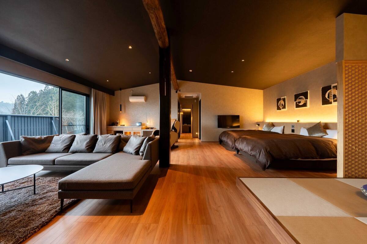 A modern hotel room with a large bed, a sofa, a dining area, and a view of the outdoors.