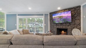 Een woonkamer met een stenen open haard, een grote flatscreen televisie en uitzicht op een huis door een glazen deur.
