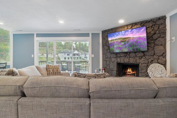 A living room with a stone fireplace, a large flat-screen TV, and a view of a house through a glass door.