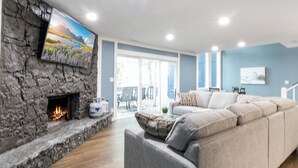 A modern living room with a stone fireplace, a large flat-screen TV, a grey sectional sofa, and a glass door leading to an outdoor area.