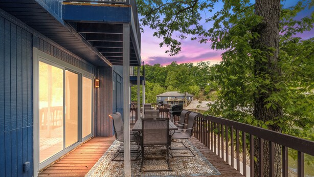 A deck with a table and chairs, overlooking a forested area with a house in the background.