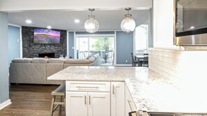 A modern kitchen with a stone fireplace, a dining area with a view, and a living room with a gray sofa.