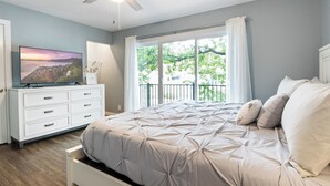 A bedroom with a bed, a TV on a dresser, a ceiling fan, and a window with a view of trees.