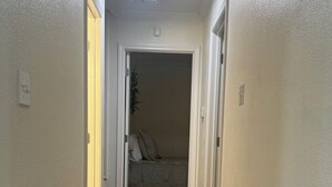 A hallway with wooden flooring, white walls, and a door leading to a room with a bed and pillows. - A Home Away From Home - Rest, Relax, & Enjoy (Alexandria)