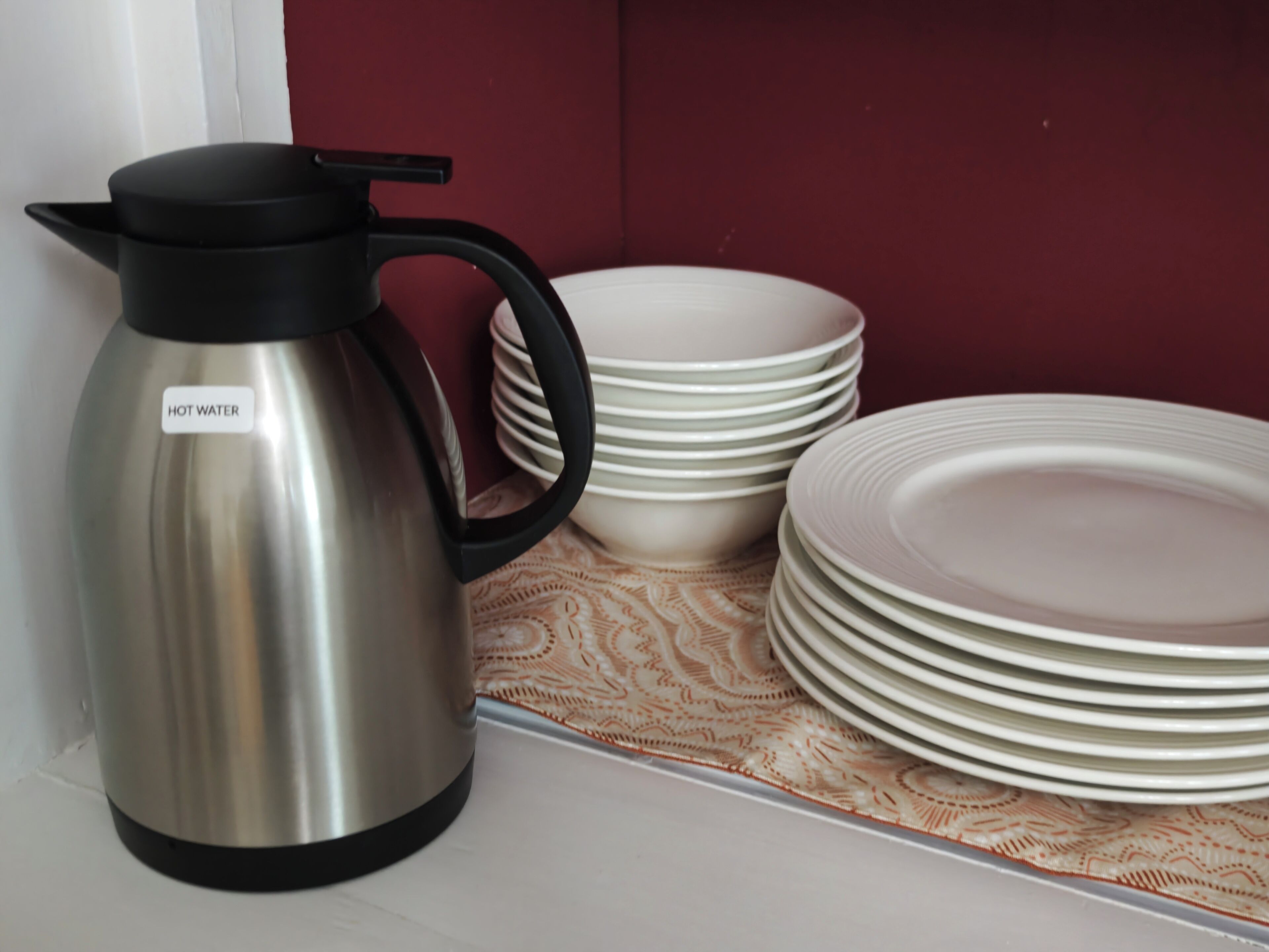 A stainless steel hot water kettle with a black handle and lid, placed on a white surface next to a stack of white plates and bowls.