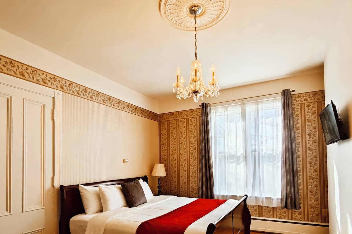 A bedroom with a large bed, a chandelier, a window with curtains, and a television on the wall.