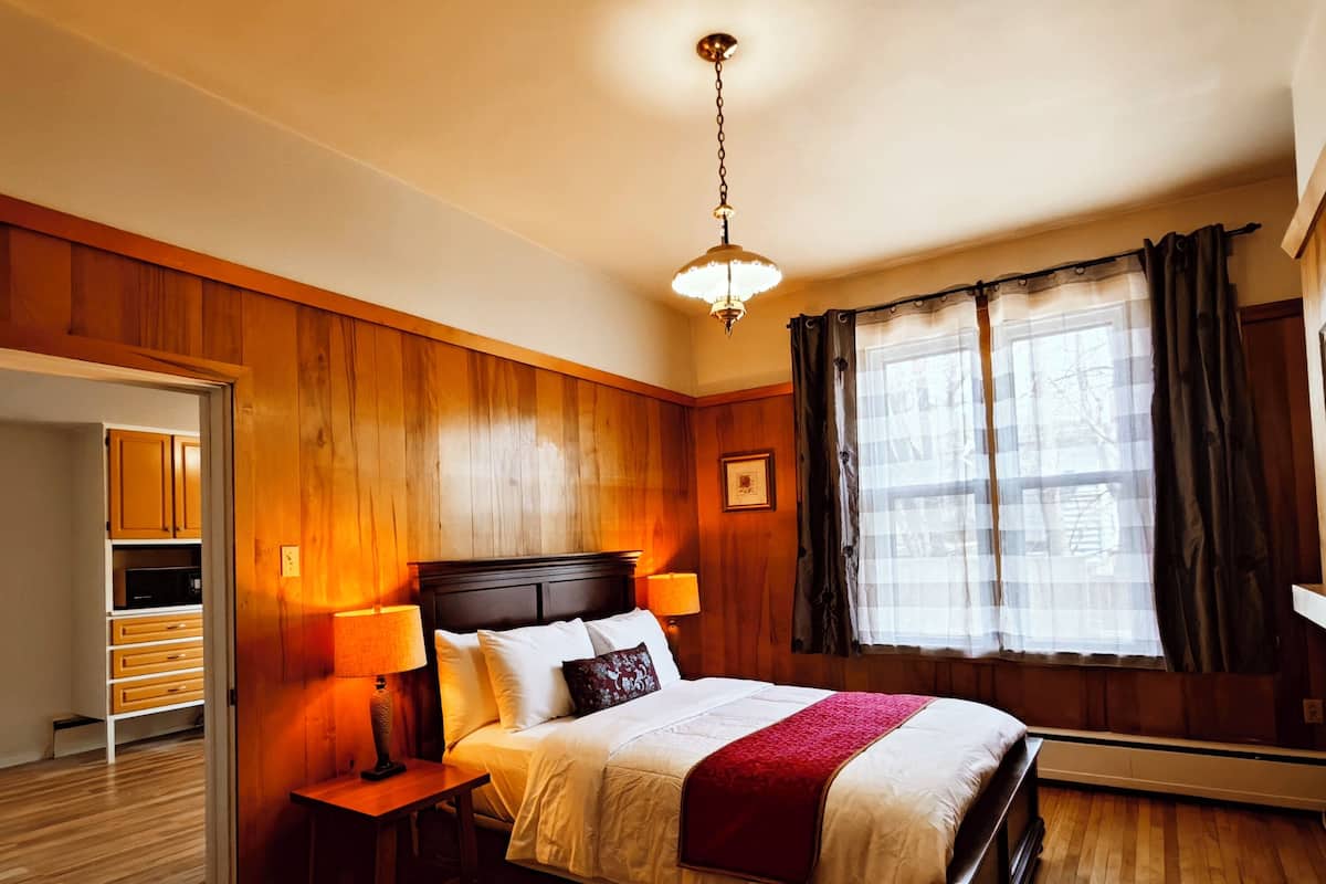 A bedroom with a wooden paneled wall, a bed with white bedding, two bedside lamps, a hanging light fixture, and a window with curtains.