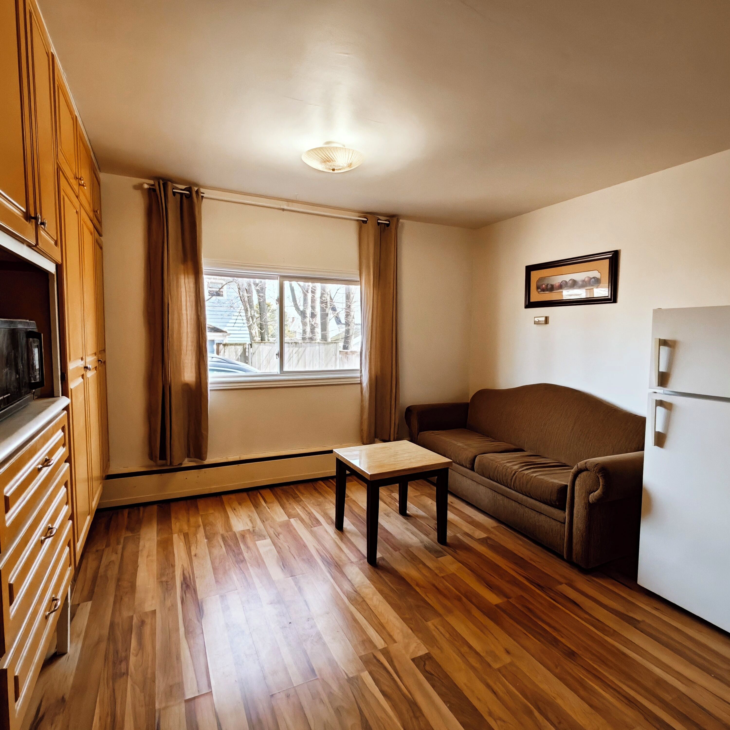 Un espace de vie compact doté d’un plancher en bois, d’un canapé, d’une petite table, d’un réfrigérateur et d’une fenêtre avec des rideaux.