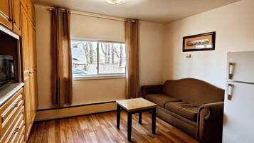 Un espace de vie compact doté d’un plancher en bois, d’un canapé, d’une petite table, d’un réfrigérateur et d’une fenêtre avec des rideaux.