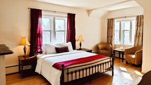 A bedroom with a large bed, two bedside tables with lamps, a chair, and two windows with curtains. - Andy's Blue House (Charlottetown)