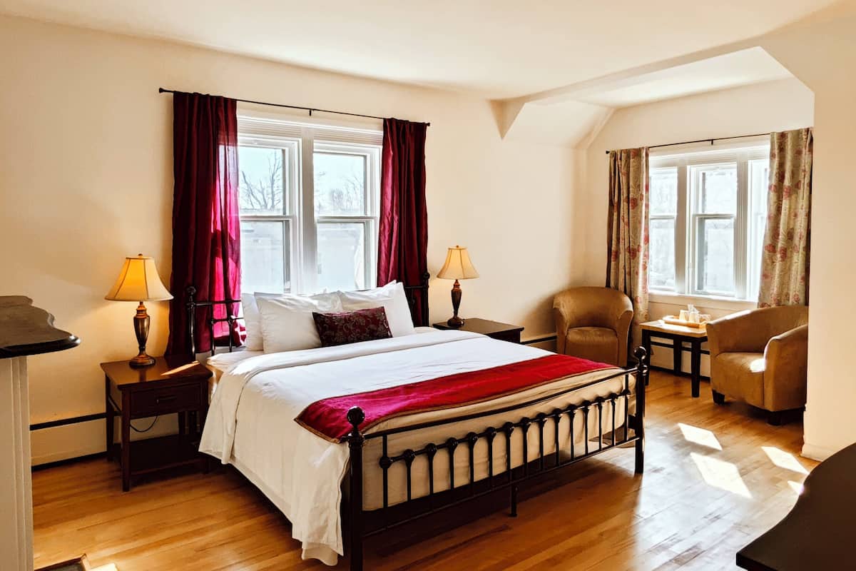 A bedroom with a large bed, two bedside tables with lamps, a chair, and two windows with curtains.