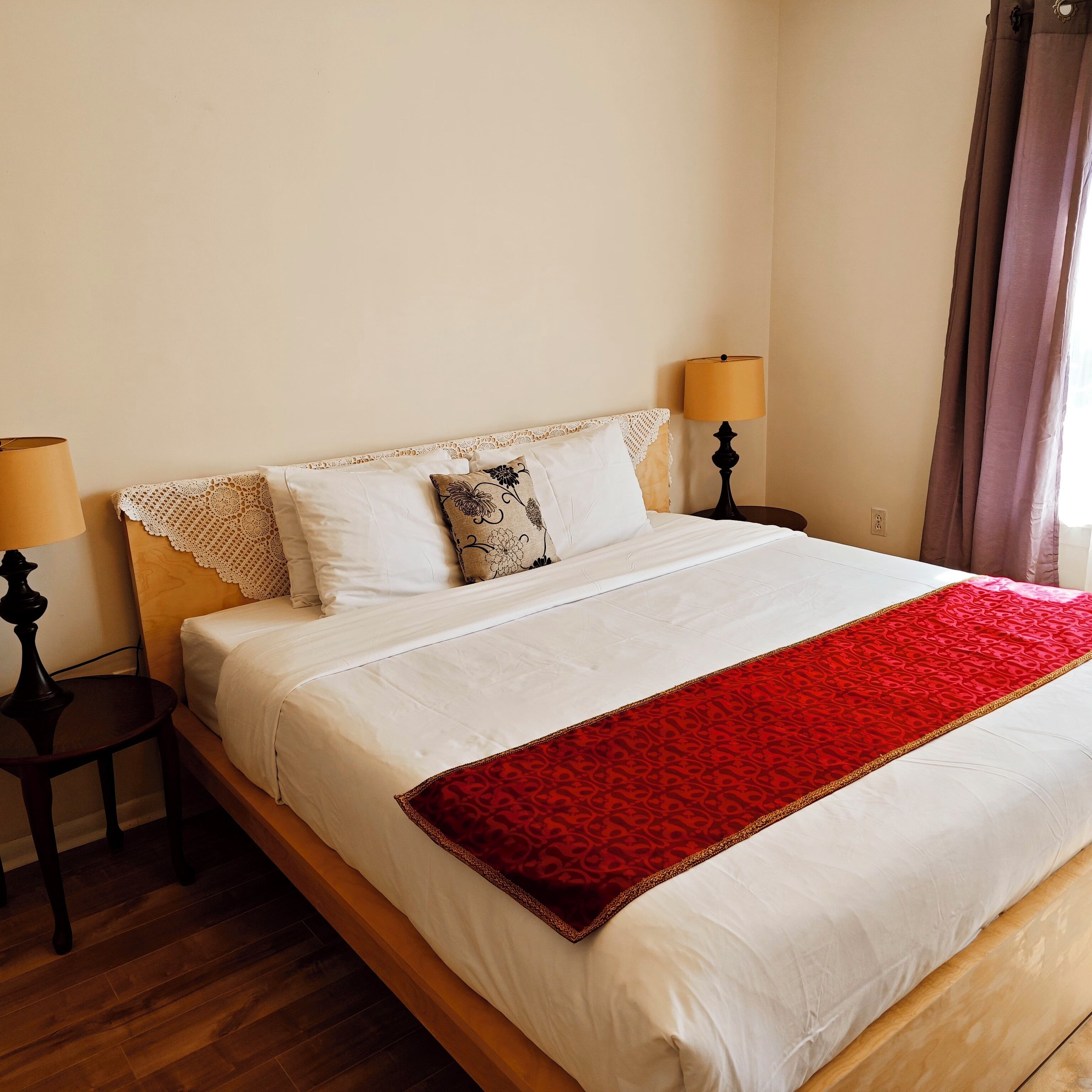 A neatly made bed with white linens and a red throw blanket, two bedside lamps, and a lace headboard.
