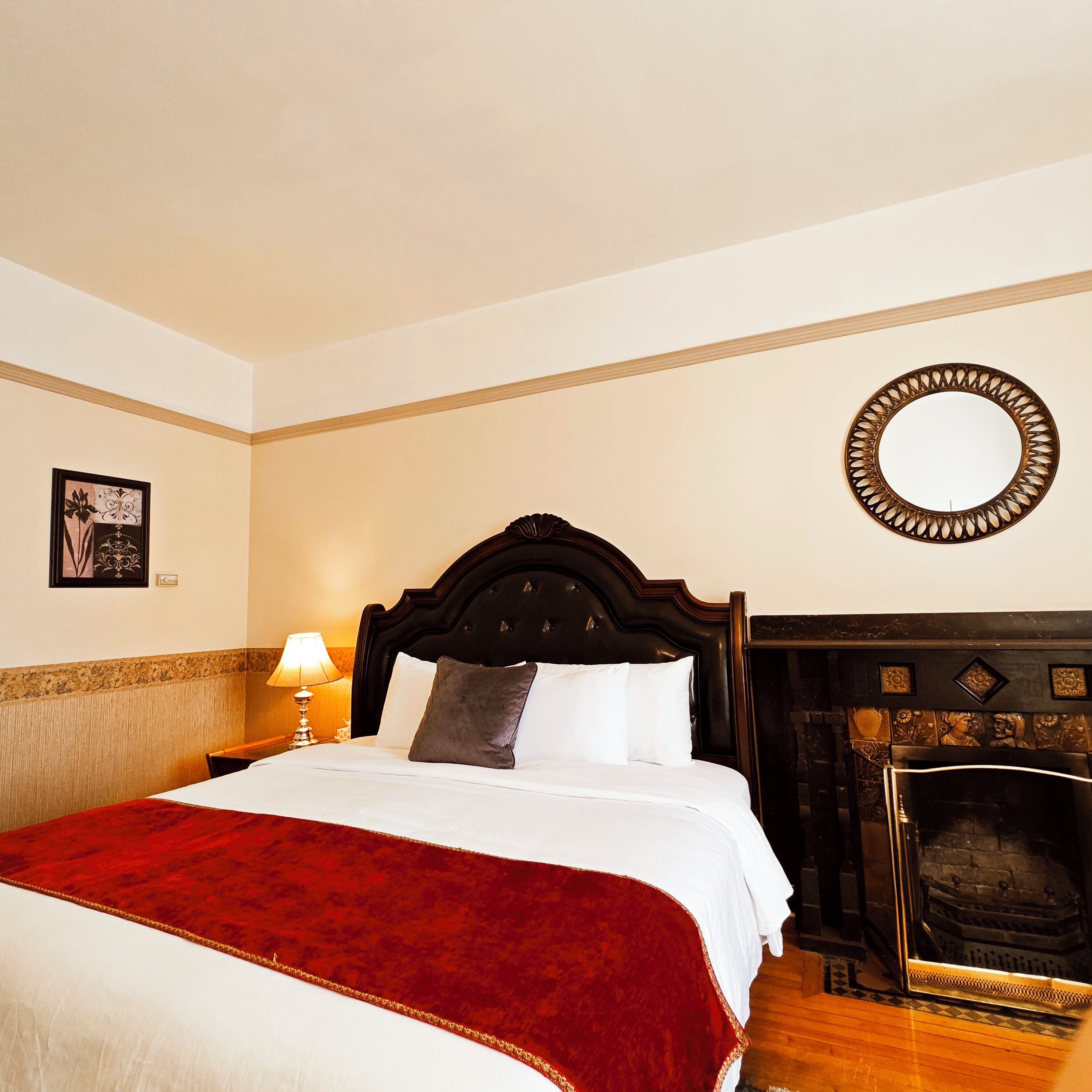 Een netjes opgemaakt bed met een rood-witte sprei, een houten hoofdbord, een wandspiegel en een open haard met decoratieve tegels.