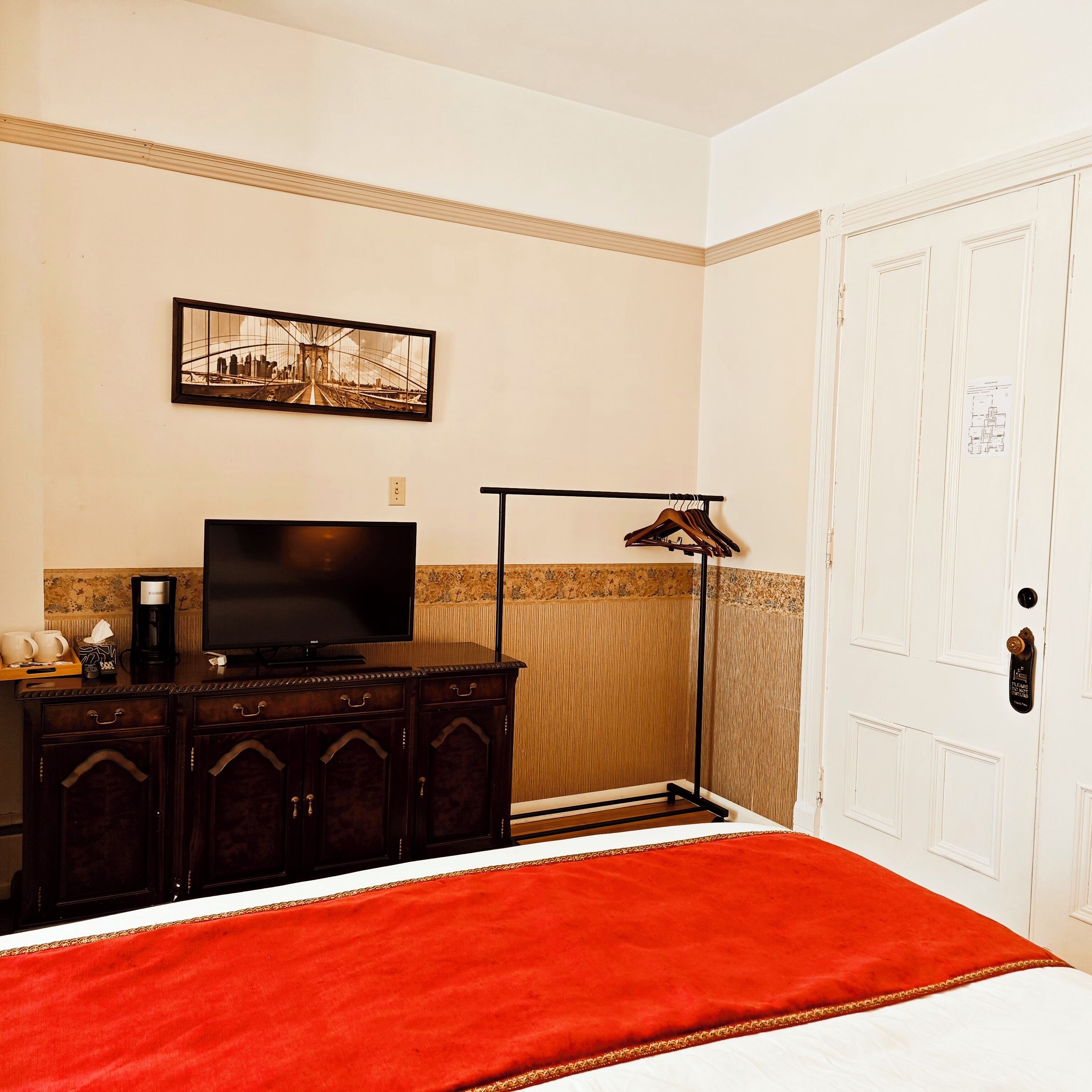 A hotel room with a bed, a TV on a wooden dresser, a coat rack, and a door with a lock.