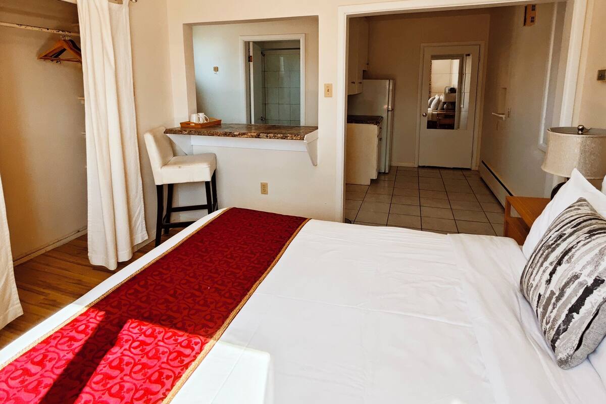 A hotel room with a bed, a desk, a chair, a wardrobe, and a kitchen area visible through an open door.