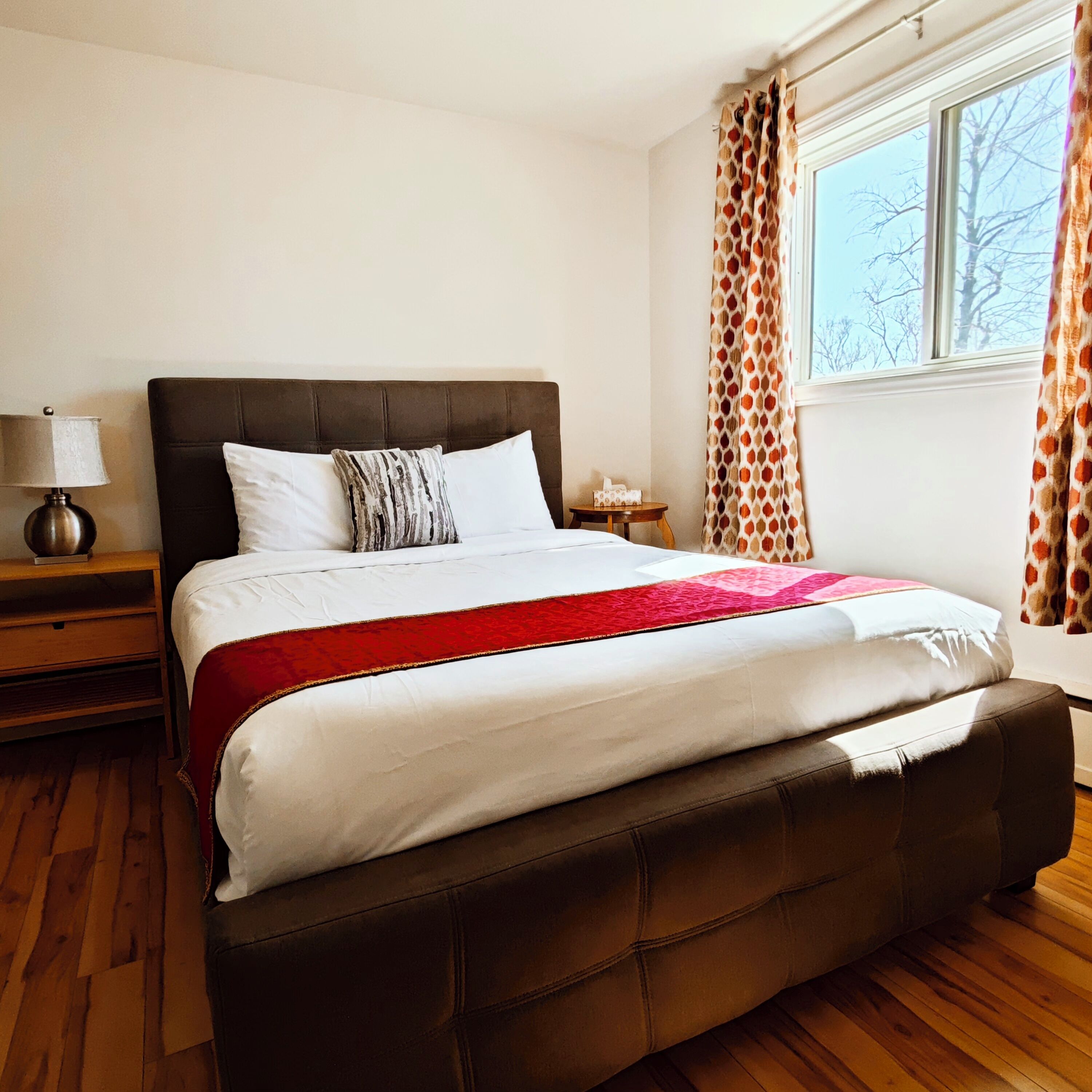A neatly made bed with a brown headboard, a bedside table with a lamp, and a window with patterned curtains.