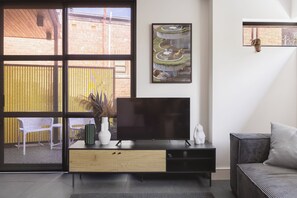 A modern living room with a large flat-screen TV on a wooden console, a wicker sofa, and a view of a brick building through a large window.