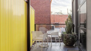 A balcony with two white chairs and a table, a yellow wall, and a brick building in the background. - MAC Middle Park by Melbourne Apartment Collection (Middle Park)