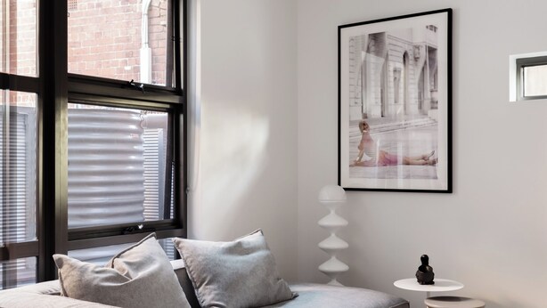 A modern living room with a grey sofa, a small round table, and a large window with blinds. - MAC Middle Park by Melbourne Apartment Collection (Middle Park)