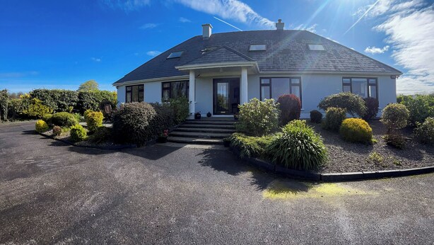 Una casa a due piani con tetto scuro, pareti esterne bianche e un portico anteriore. Diverse siepi curate e un piccolo giardino adornano la facciata.