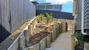 A concrete pathway leading to a building with a modern design, surrounded by wooden planters and small trees. - The King's Keep (Cable Bay)