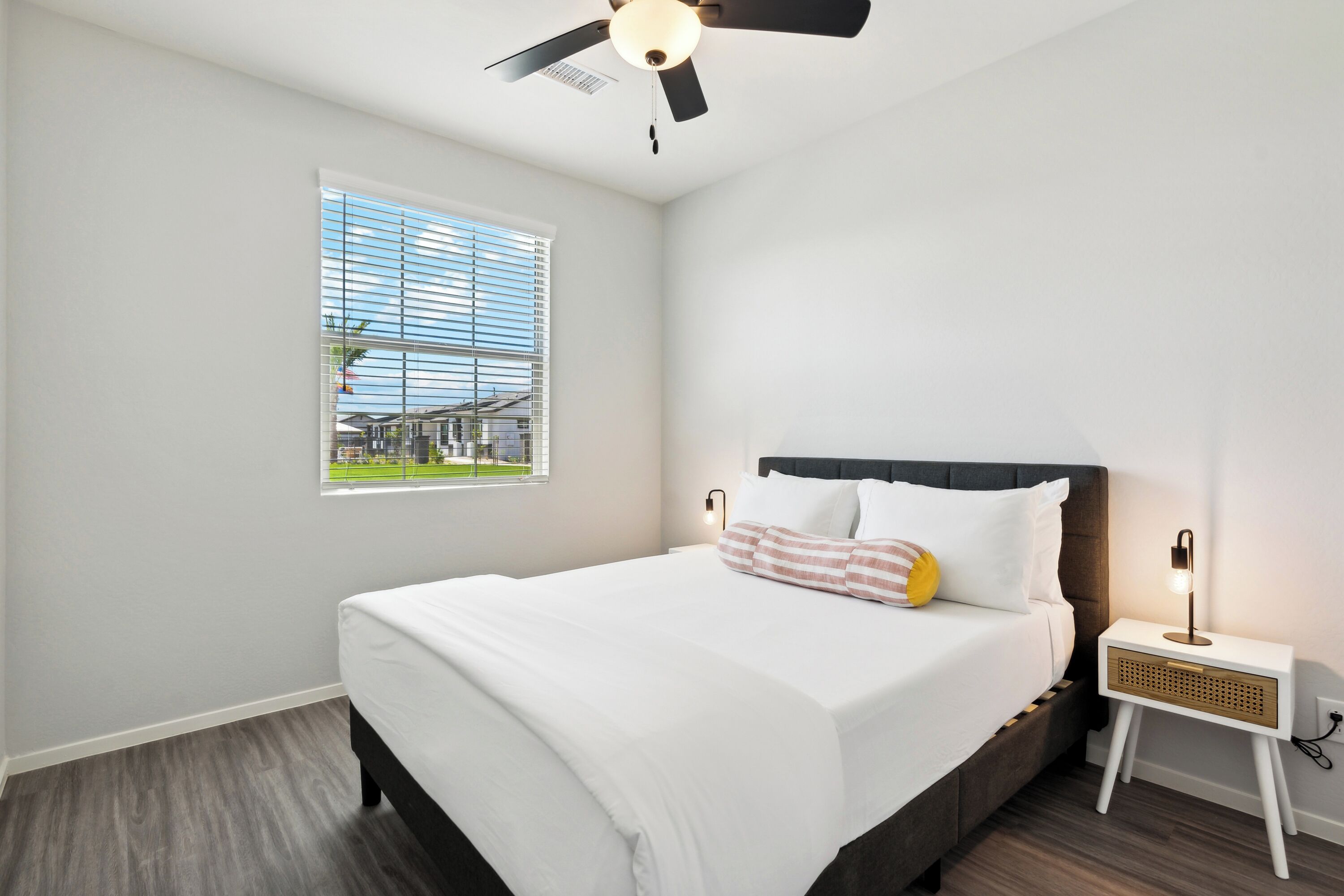 Une chambre à coucher avec un lit, une fenêtre avec des stores, un ventilateur de plafond et une table de chevet avec une lampe.