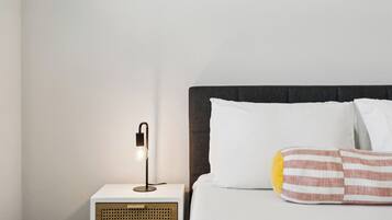 Un lit bien fait, avec des oreillers blancs et rayés, une lampe de chevet et une petite table de nuit blanche dotée d’un panneau en bois.