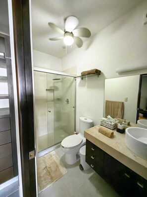 Ein modernes Badezimmer mit einer Duschkabine, einer Toilette, einem Waschtisch mit großem Becken und einem Deckenventilator.