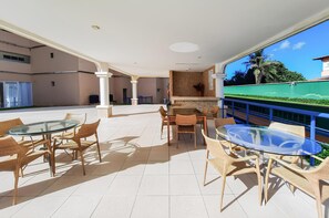 Une vaste terrasse extérieure dotée d’une table en verre et de chaises en osier, entourée d’un sol carrelé blanc et d’un petit muret en briques.
