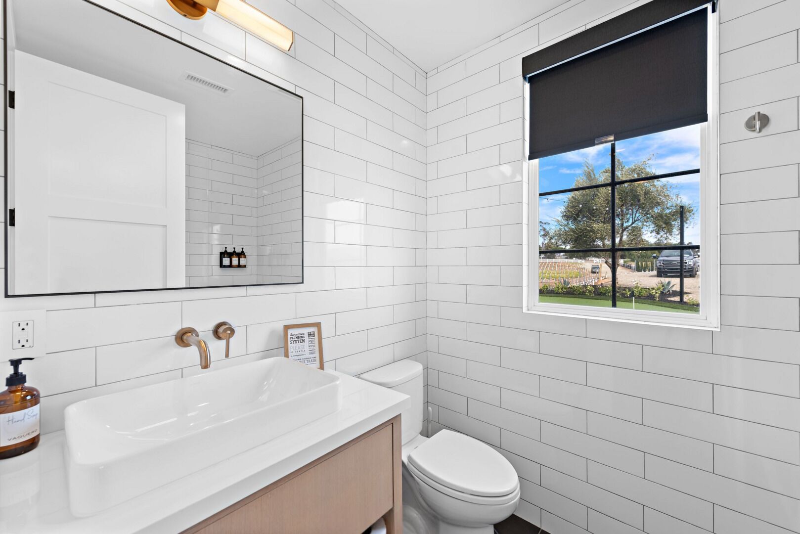 A modern bathroom with white subway tiles, a large mirror, a white sink, a toilet, and a window with a view of the outside.