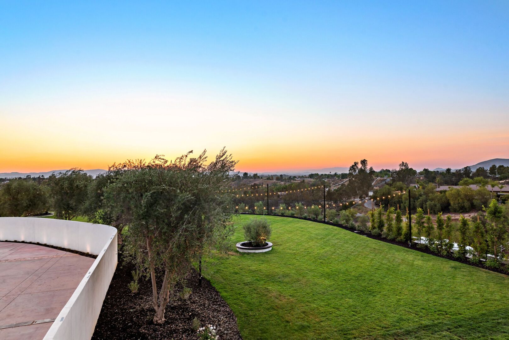 A landscaped garden with a curved pathway, a circular planter, and a view of a residential area during sunset.