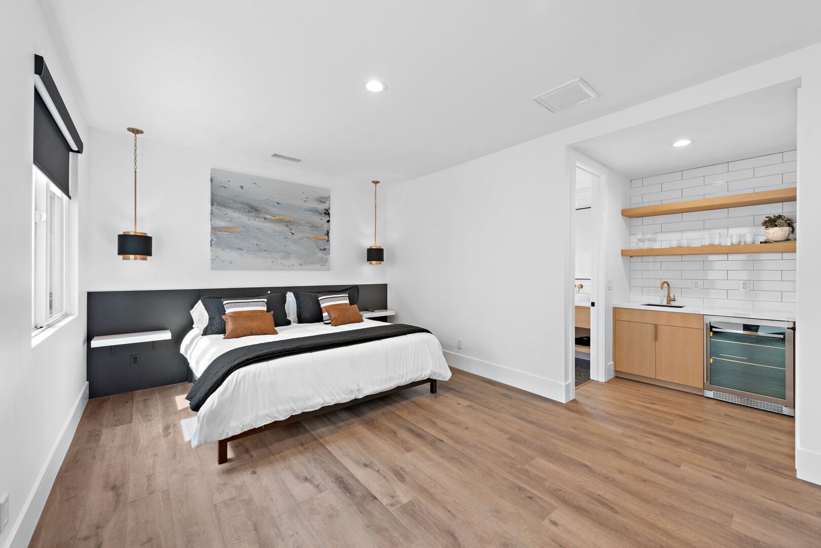 A modern bedroom with a large bed, a bedside table, and a view into the kitchen area.
