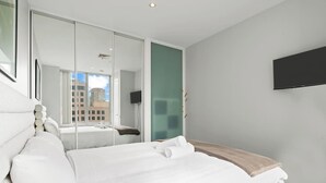 A modern hotel room with a large bed, a glass partition, a television, and a view of the cityscape.