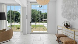 A modern interior with a large glass door leading to a garden, a wicker sofa, a console table with a vase, and a hanging light fixture. - Oceanview Oasis: Luxury 6BR Home with Pool (Seagrove Beach)
