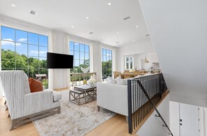 A spacious living room with a large window, a flat-screen TV, a sofa, a coffee table, and a staircase leading to an upper level. - Oceanview Oasis - 6BR Seagrove Beach Home on 30A - Private Pool - Sleeps 12 (Seagrove Beach)