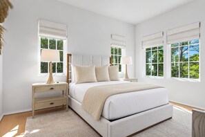 Une chambre à coucher avec un grand lit, deux tables de chevet et trois fenêtres équipées de stores blancs.