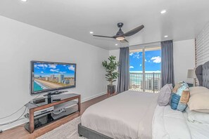 A modern bedroom with a large bed, a television on a wooden stand, a ceiling fan, and a view of the ocean through a balcony. - Holiday Towers 1503 (Myrtle Beach)