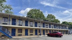 Un motel amb una filera d'habitacions, cadascuna amb una porta blava i un balcó. Hi ha un aparcament amb un cotxe vermell aparcat al davant.