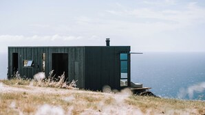 Eine moderne, dunkel gefärbte Hütte mit einem Eingang und einer kleinen Veranda, gelegen auf einem grasbewachsenen Hügel mit Blick auf ein Gewässer.