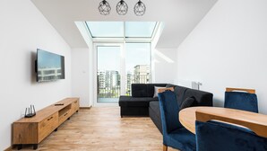 A modern living room with a black sectional sofa, a wooden dining table, and blue chairs. A TV is mounted on the wall, and there is a wooden cabinet with drawers.