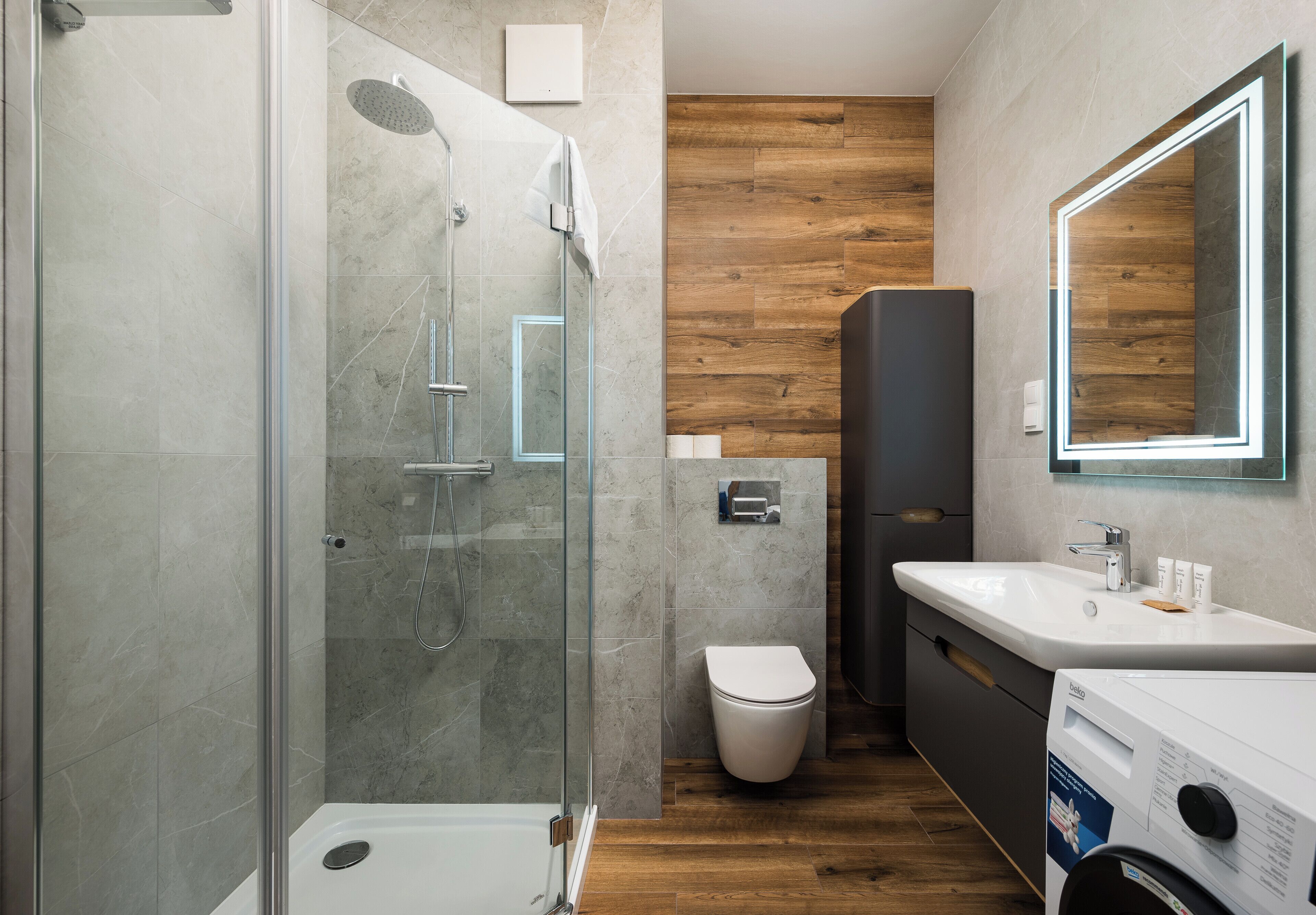 Un baño moderno con ducha de vidrio, piso de madera, inodoro blanco, lavamanos con espejo y lavadora.