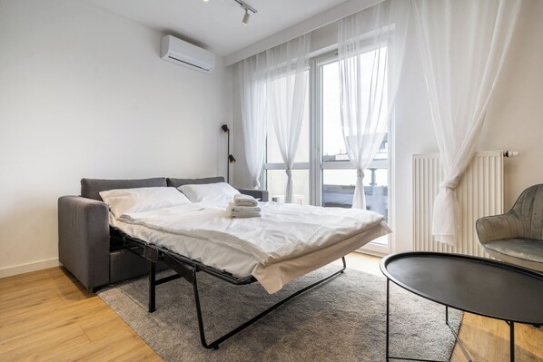 A modern hotel room with a bed, a small table, a chair, and a window with sheer curtains.