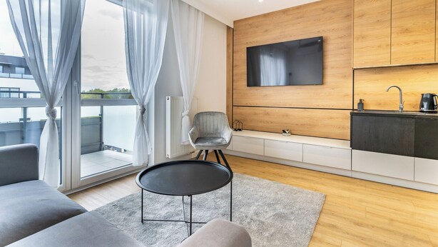 Un salon moderne comprenant un canapé, une petite table, un fauteuil, un coin cuisine avec un évier et une machine à café, ainsi qu’un téléviseur fixé au mur.