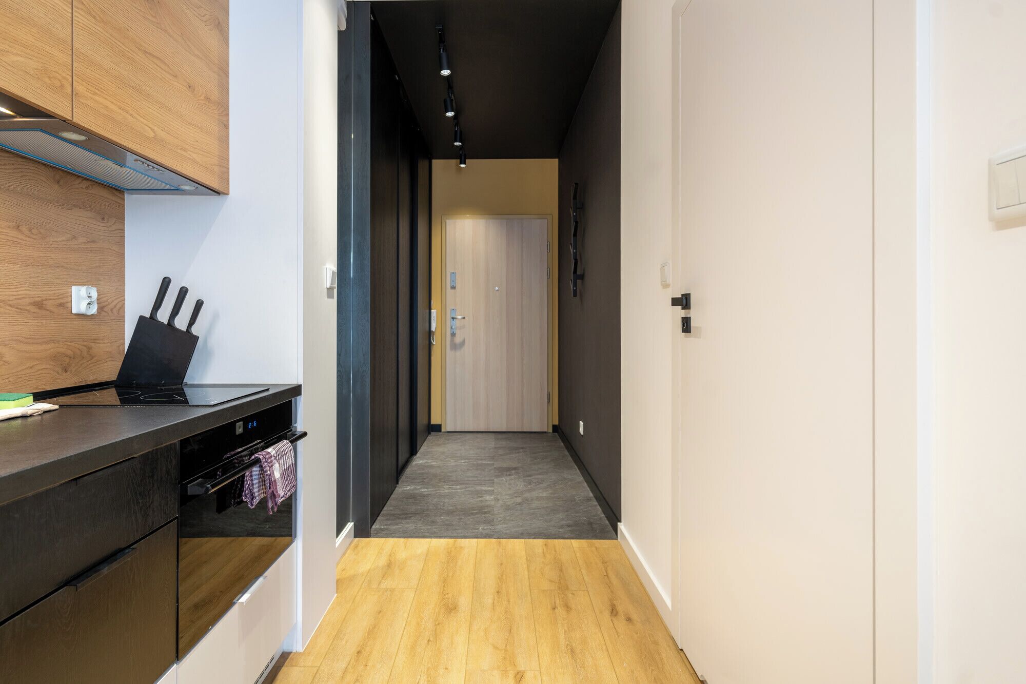 Un pasillo estrecho con piso de madera, una zona de cocina con cubierta negra y un bloque de cuchillos, y una puerta que da a otra habitación.