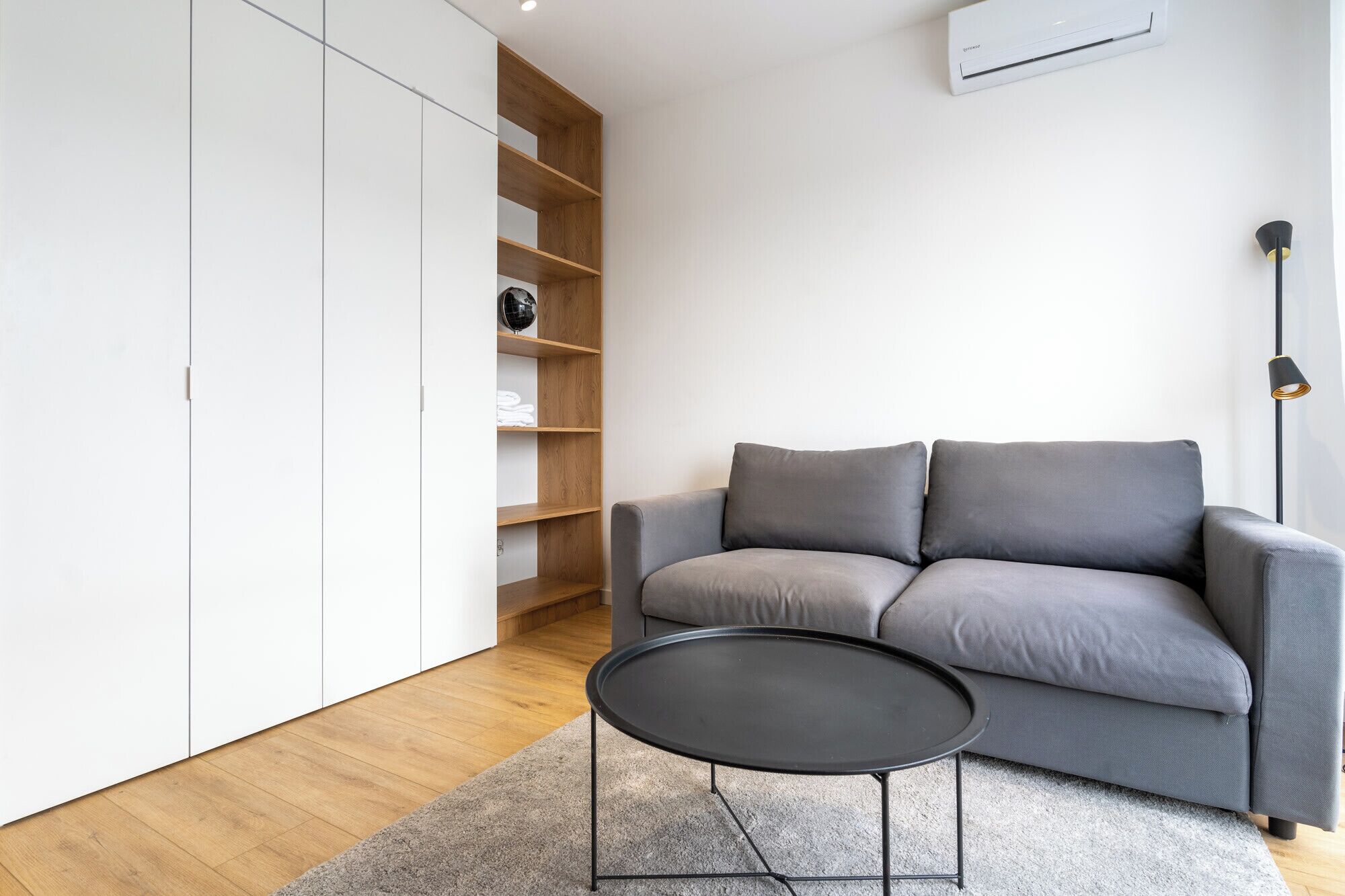 Una sala moderna con un sofá gris, una mesa de centro negra, una estantería de madera y una lámpara de pie.