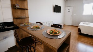 A modern kitchen with a wooden table set for two, a TV mounted on the wall, and a white sofa in the living area. - Olímpia Aparts  (Olímpia)